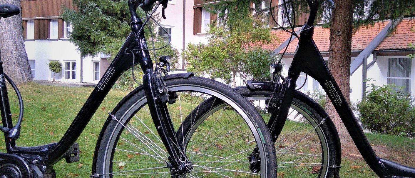 Two black e-bikes are parked on a green lawn in front of a multi-storey building with wooden cladding and windows., &copy; relexa Waldhotel Schatten