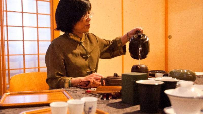 Eine Frau in traditioneller Kleidung gie&szlig;t Tee in einer chinesischen Teeschule ein. Der Raum ist mit Holzw&auml;nden und Teezubeh&ouml;r dekoriert., &copy; Bambusbr&uuml;cke Stuttgart
