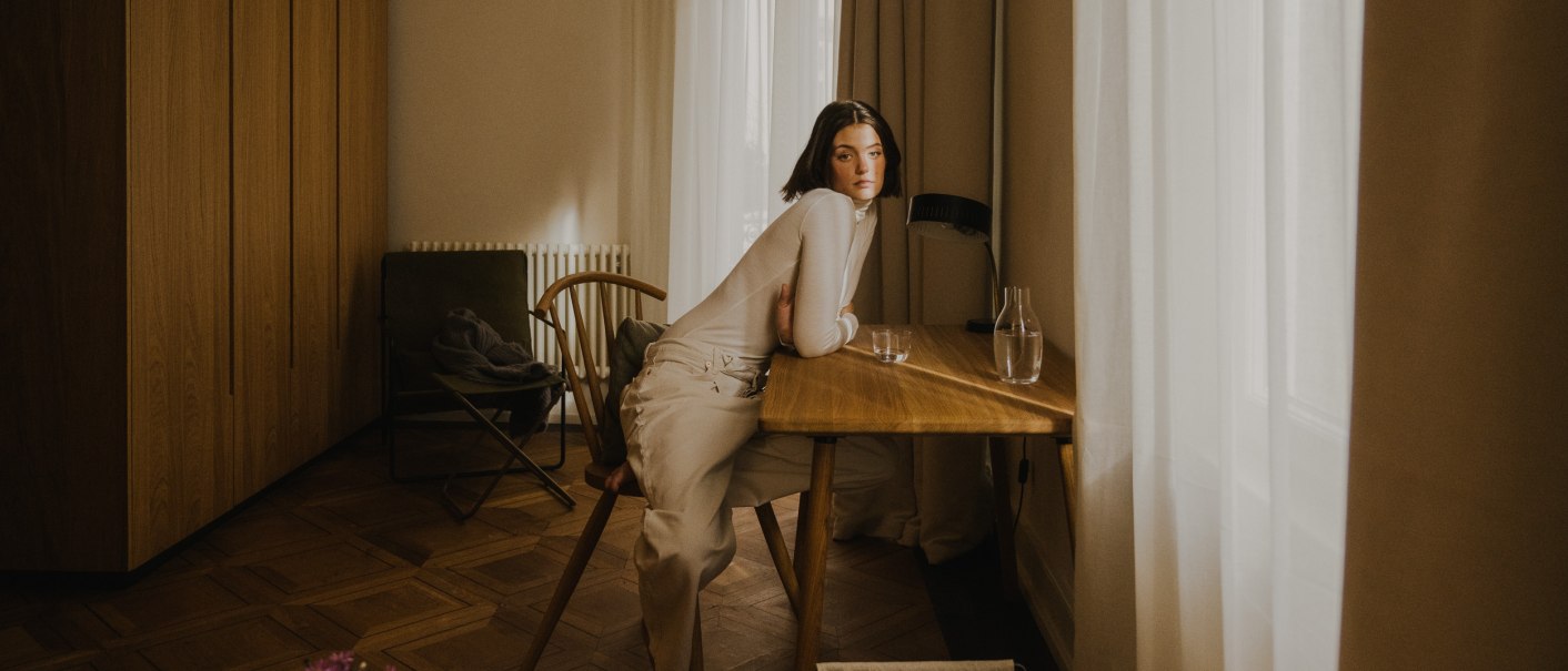 Frau in wei&szlig;er Kleidung sitzt an einem Holztisch in einem stilvoll eingerichteten Raum mit Vorh&auml;ngen und Holzboden., &copy; Peter Oliver Wolff / DAS EDITH