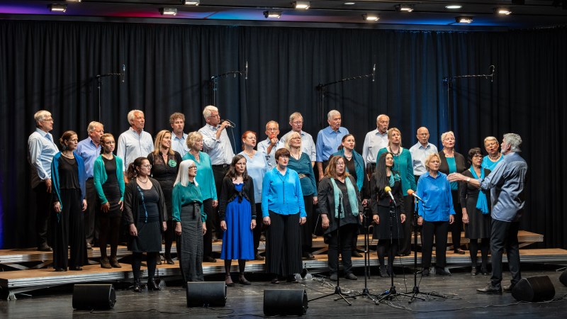 Ein Chor in blauen und schwarzen Outfits singt auf einer Bühne. Der Dirigent steht rechts und leitet die Gruppe an. Mikrofone sind aufgestellt., © Filip Filipov / OnnenChor
