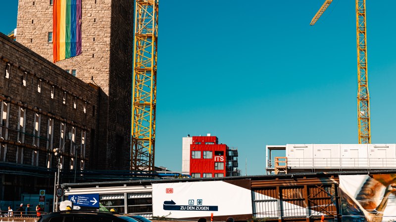 Ein Gebäude mit Regenbogenfahne, daneben ein gelber Baukran. Im Hintergrund ein rotes Gebäude mit der Aufschrift 'ITS'. Ein Schild weist zu den Zügen., © Stuttgart Marketing GmbH, Sarah Schmid Ein Gebäude mit Regenbogenfahne, daneben ein gelber Baukran. Im Hintergrund ein rotes Gebäude mit der Aufschrift 'ITS'. Ein Schild weist zu den Zügen., © Stuttgart Marketing GmbH, Sarah Schmid