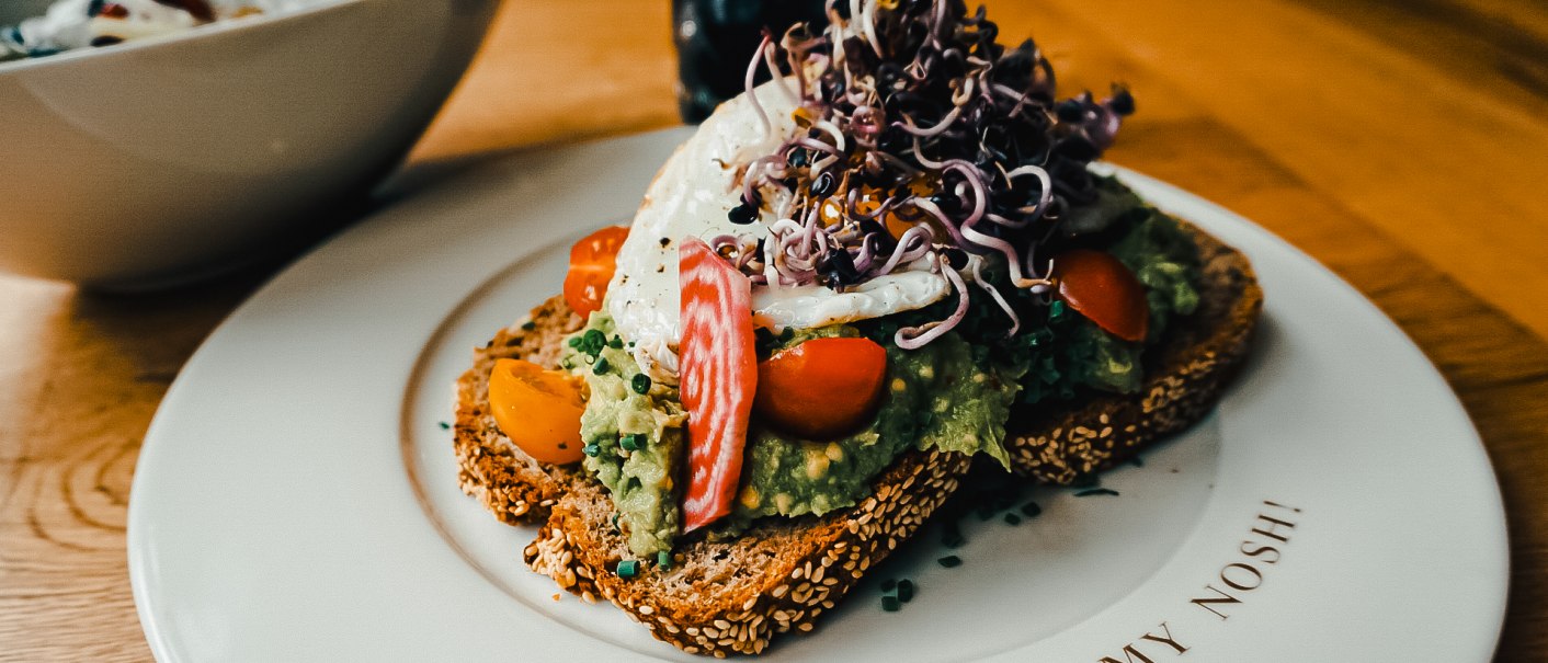 Belegtes Brot mit Avocado, Tomaten und Sprossen auf einem Teller mit der Aufschrift 'Oh my Nosh'., © The Gardeners Nosh