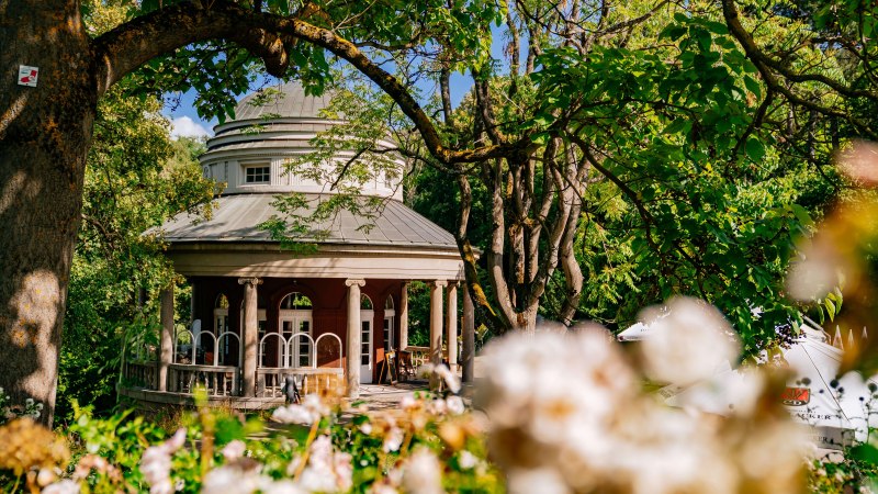 A round teahouse with pillars in Weissenburg Park, surrounded by green trees and blooming flowers in the foreground., © Thomas Niedermüller