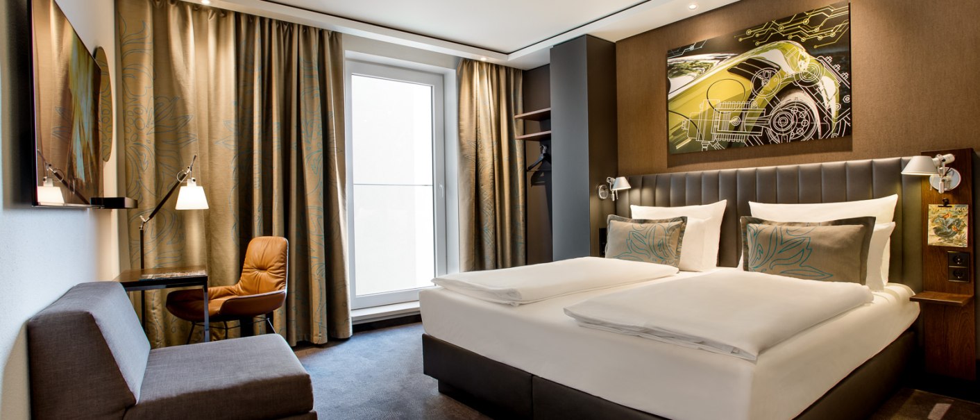 Modernes Hotelzimmer mit Doppelbett, Sofa, Schreibtisch und großem Fenster. An der Wand hängt ein Kunstwerk mit einem Auto-Motiv., © TOMAS
