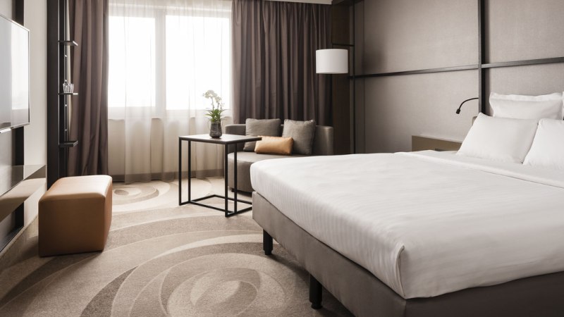 Modernes Hotelzimmer mit großem Bett, Sofa, Tisch und Fernseher. Helle Vorhänge und dezente Farben schaffen eine elegante Atmosphäre., © Stuttgart Marriott Hotel Sindelfingen