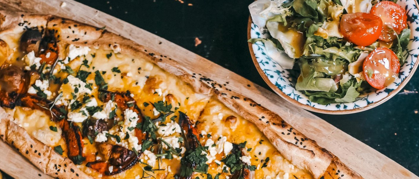 A flatbread topped with cheese and vegetables on a wooden board, next to it a fresh salad with tomatoes in a bowl., © Noa, Stuttgart