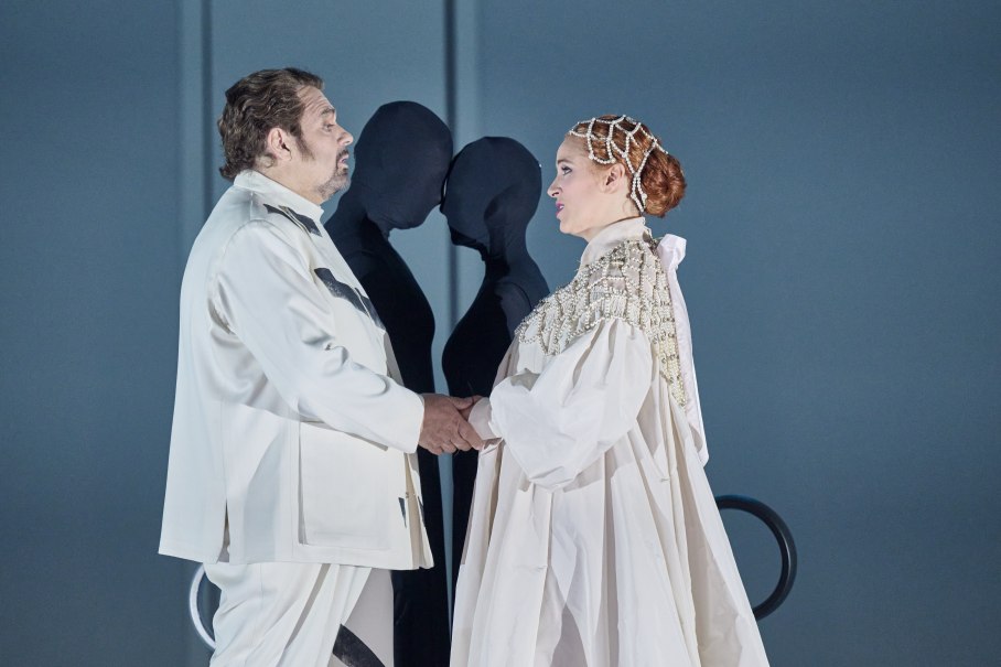 Zwei Opernsänger in weißen Kostümen stehen sich gegenüber und halten sich die Hände. Im Hintergrund sind zwei schwarze Figuren zu sehen., © Martin Sigmund