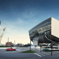 The Porsche Museum in Stuttgart with its futuristic architecture and a sculpture of three cars on tall pillars against a blue sky., &copy; Porsche AG