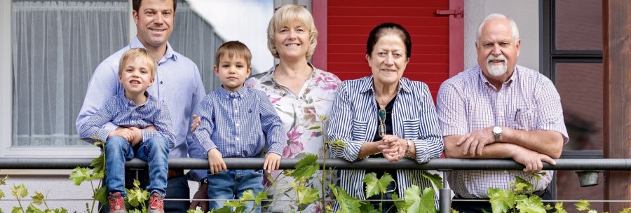 A smiling family stands on a balcony. Two children sit on the railing, surrounded by adults. Green plants can be seen in the foreground., © Weingut Zaiß