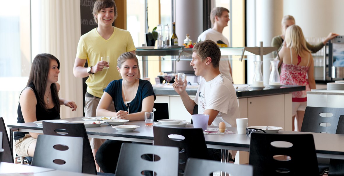 Young people are sitting and standing in a modern dining room, chatting and drinking. In the background, a counter with dishes is visible., © DJH Landesverband Baden-Württemberg e.V. Young people are sitting and standing in a modern dining room, chatting and drinking. In the background, a counter with dishes is visible., © DJH Landesverband Baden-Württemberg e.V.