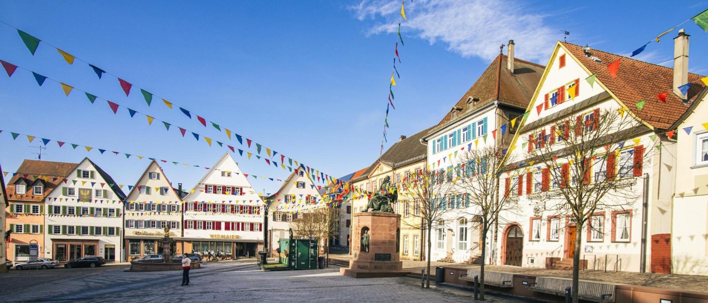The town square in Weil der Stadt features historic buildings and colorful pennants against a blue sky. A statue stands in the center of the square., © SMG Stuttgart Marketing GmbH - Sarah Schmid The town square in Weil der Stadt features historic buildings and colorful pennants against a blue sky. A statue stands in the center of the square., © SMG Stuttgart Marketing GmbH - Sarah Schmid