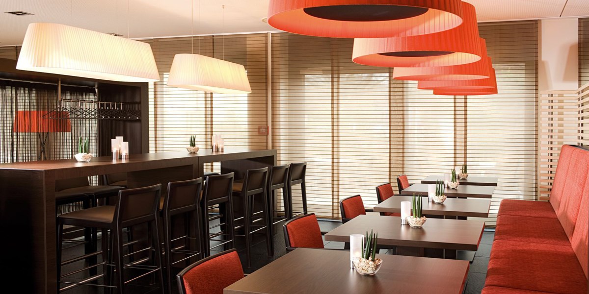 A modern breakfast room with red lamps, wooden tables, and chairs. Large windows with blinds let in plenty of light., © NH Ludwigsburg A modern breakfast room with red lamps, wooden tables, and chairs. Large windows with blinds let in plenty of light., © NH Ludwigsburg
