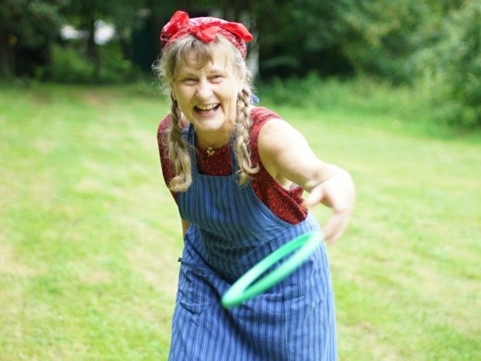 Frau mit Zöpfen und rotem Kopftuch wirft einen grünen Ring auf einer Wiese. Sie trägt ein blau gestreiftes Kleid und lächelt fröhlich., © Cool-Tours StattReisen Frau mit Zöpfen und rotem Kopftuch wirft einen grünen Ring auf einer Wiese. Sie trägt ein blau gestreiftes Kleid und lächelt fröhlich., © Cool-Tours StattReisen