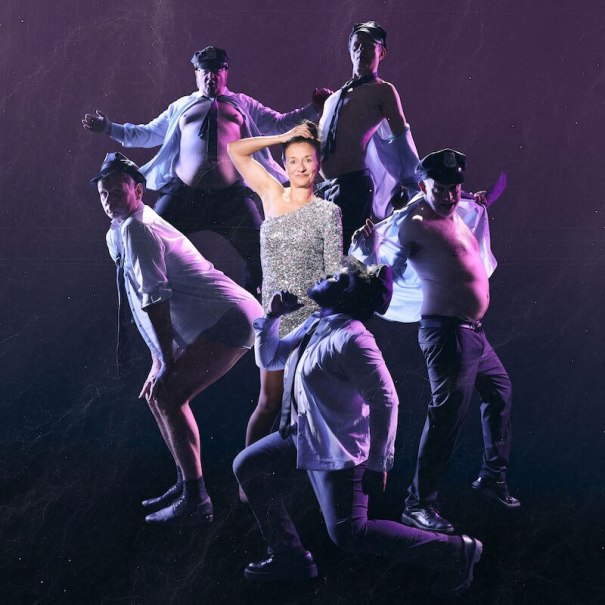 A woman in a glittering dress poses with five men in police uniforms in front of a purple background. The men dance and pose in various poses., © Theaterhaus Stuttgart e.V. A woman in a glittering dress poses with five men in police uniforms in front of a purple background. The men dance and pose in various poses., © Theaterhaus Stuttgart e.V.