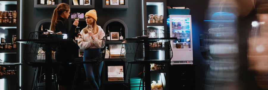 Zwei Frauen in einem stilvollen Café, eine mit gelber Mütze, halten Weingläser. Im Hintergrund sind Regale mit Produkten und ein Kühlschrank zu sehen., © Stuttgart-Marketing GmbH, Alwin Maigler