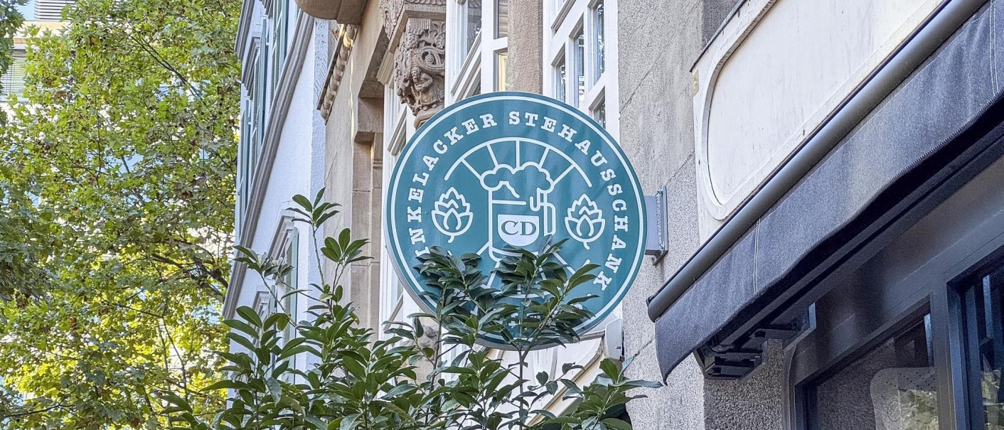 Schild von WELLERS Dinkelacker Stehausschank an einer Hausfassade, umgeben von Grün und Stadtarchitektur., © SMG, Ellen Haeffner Schild von WELLERS Dinkelacker Stehausschank an einer Hausfassade, umgeben von Grün und Stadtarchitektur., © SMG, Ellen Haeffner