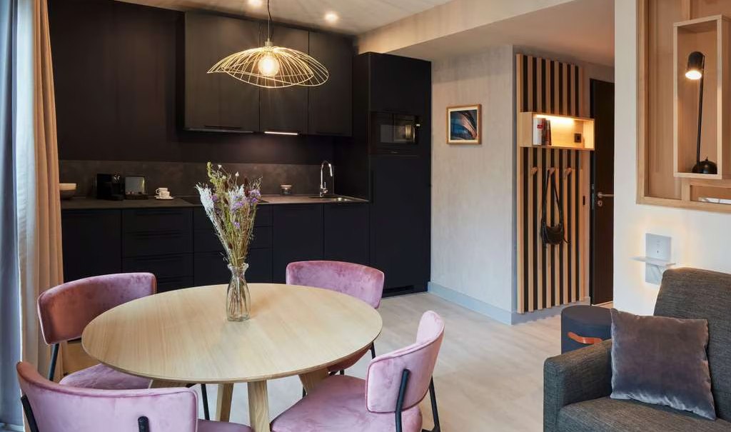 Modernes Apartment mit rundem Holztisch, rosa Stühlen, dunkler Küche und stilvoller Beleuchtung. Gemütliche Atmosphäre mit Sofa und Dekoration., © Adagio