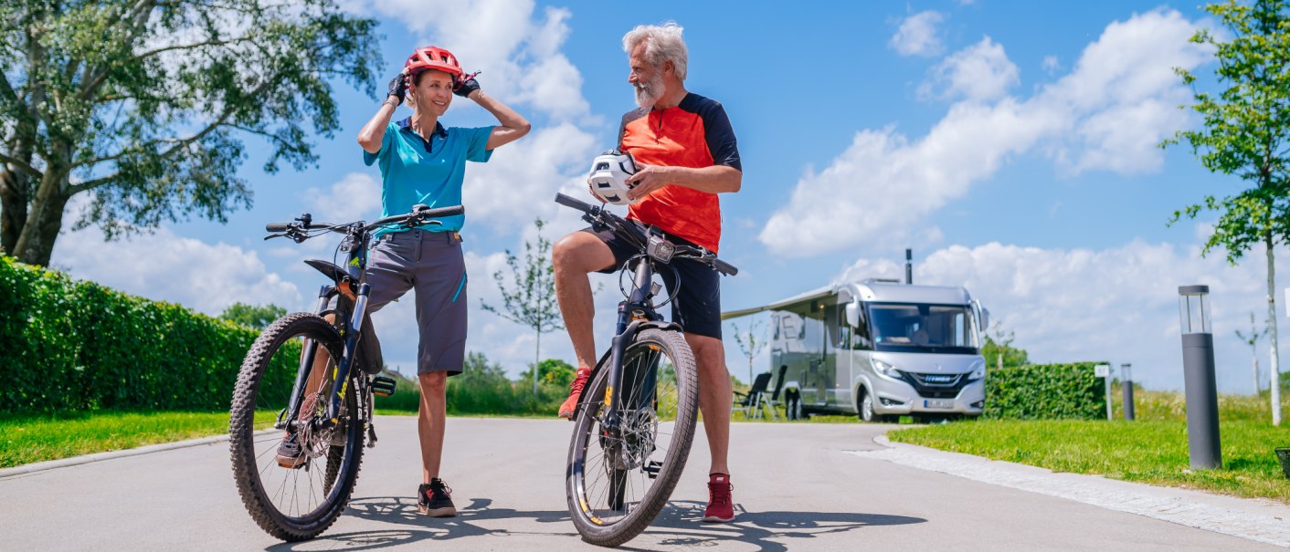 Ein älteres Paar mit Fahrrädern genießt einen sonnigen Tag auf einer Straße. Im Hintergrund steht ein modernes Wohnmobil., © Thomas Niedermüller Ein älteres Paar mit Fahrrädern genießt einen sonnigen Tag auf einer Straße. Im Hintergrund steht ein modernes Wohnmobil., © Thomas Niedermüller