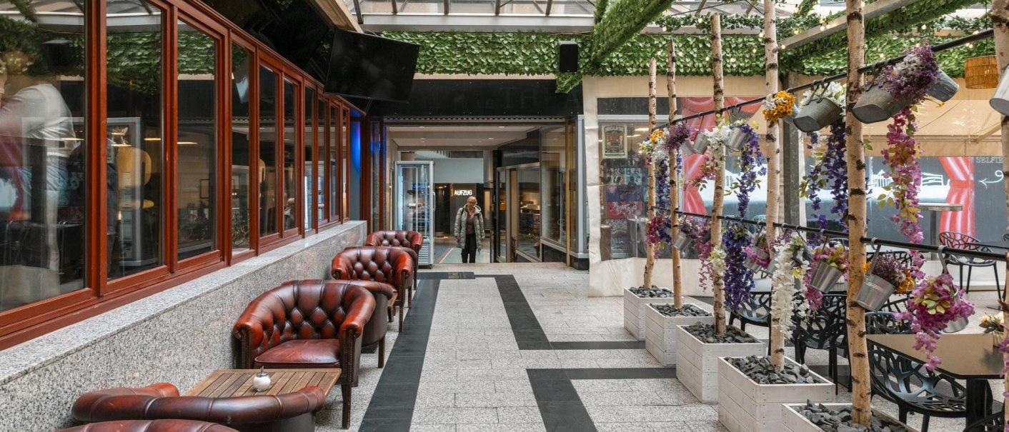 Ein gem&uuml;tlicher Innenhof mit roten Ledersesseln, dekorativen Pflanzen und einem Glasdach. Eine Person betritt den Bereich im Hintergrund., &copy; SMG, Sarah Schmid