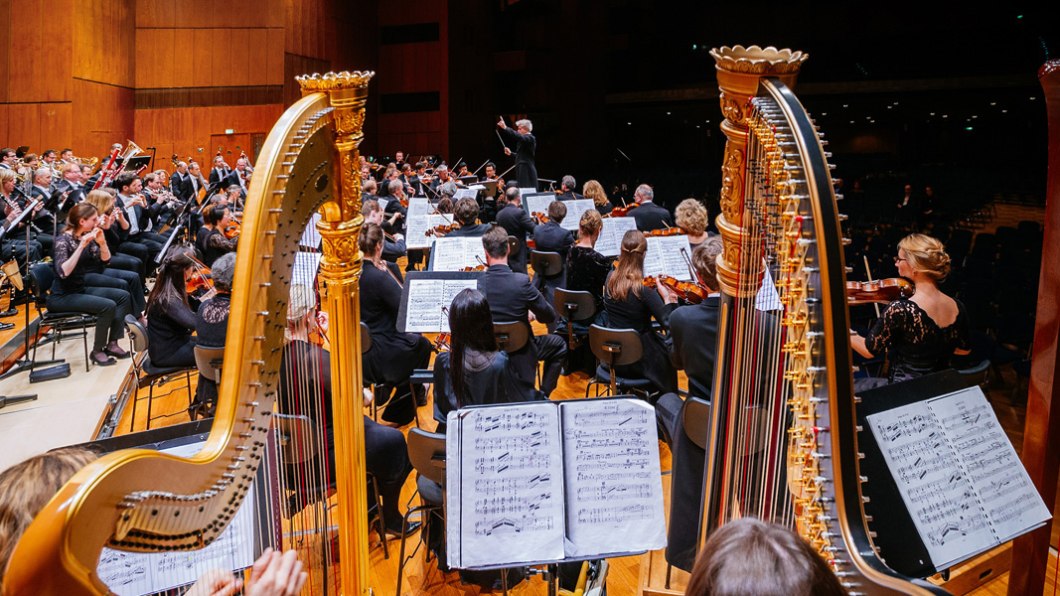 Ein Orchester spielt auf einer Bühne, mit zwei Harfen im Vordergrund. Der Dirigent steht vor den Musikern, die Notenblätter lesen., © Stuttgarter Philharmoniker Ein Orchester spielt auf einer Bühne, mit zwei Harfen im Vordergrund. Der Dirigent steht vor den Musikern, die Notenblätter lesen., © Stuttgarter Philharmoniker