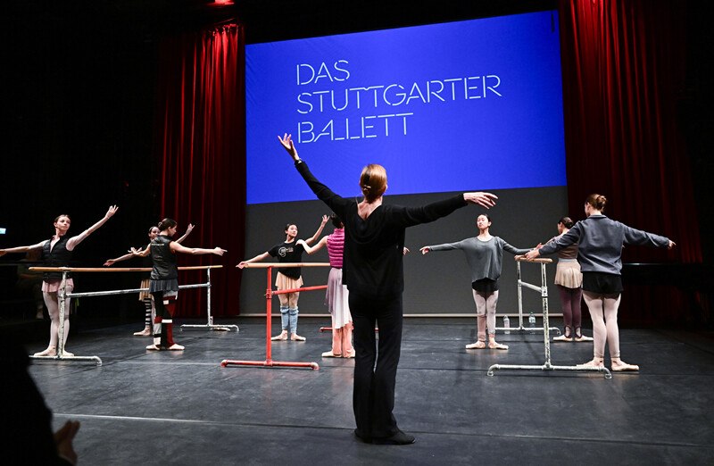 Ballettprobe mit T&auml;nzern und Trainerin vor einer Leinwand mit der Aufschrift 'Das Stuttgarter Ballett'., &copy; W&uuml;rttembergische Staatstheater Stuttgart