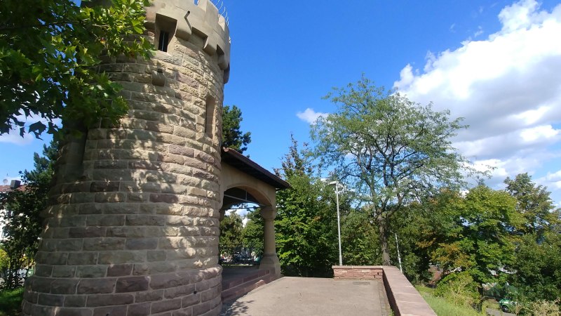Runder Steinturm mit Zinnen, umgeben von B&auml;umen und blauem Himmel. Der Turm steht auf einer gepflasterten Fl&auml;che., &copy; SMG