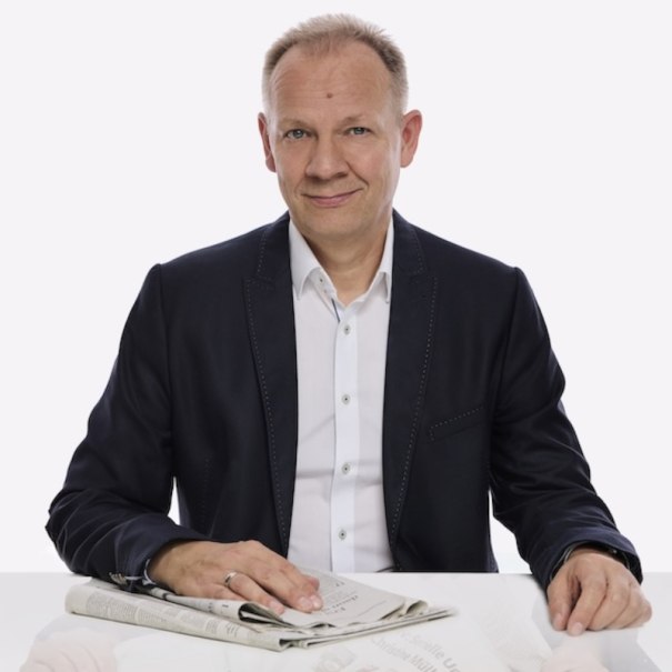 A man in a dark suit is sitting at a table holding a newspaper. The background is white., &copy; Renitenztheater Stuttgart e.V.