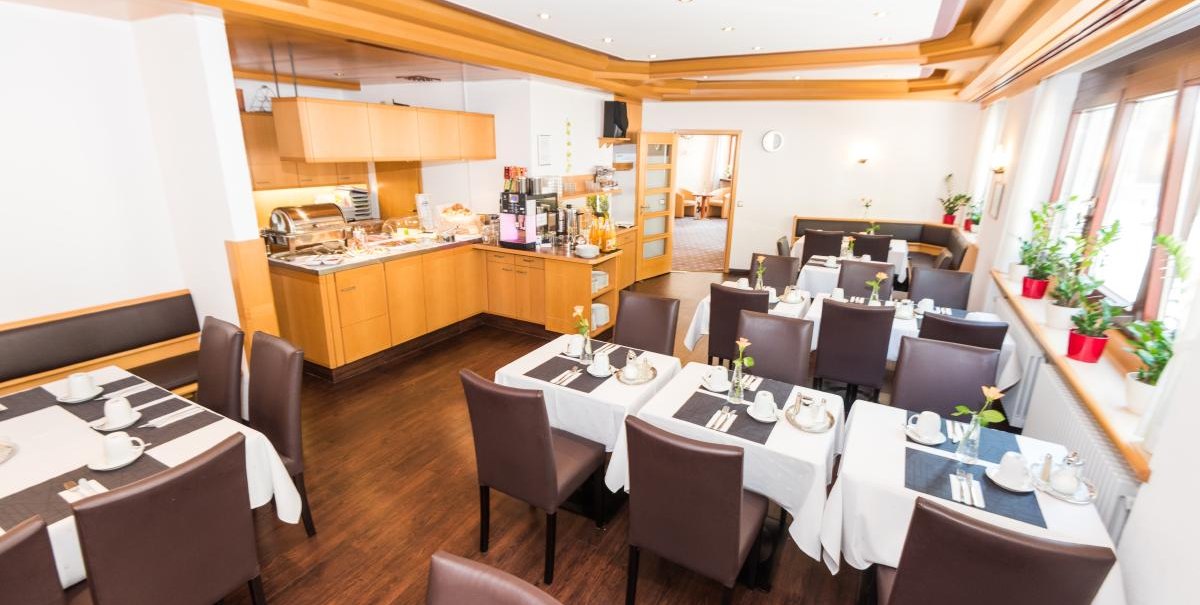 A bright breakfast room with laid tables, brown chairs and a buffet area. There are plants on the windowsill., © ARNOTEL GmbH