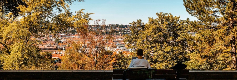 Eine Person sitzt auf einer Bank und schaut auf eine Stadt, umgeben von herbstlichen Bäumen. Der Himmel ist klar und blau., © Stuttgart Marketing GmbH, Sarah Schmid Eine Person sitzt auf einer Bank und schaut auf eine Stadt, umgeben von herbstlichen Bäumen. Der Himmel ist klar und blau., © Stuttgart Marketing GmbH, Sarah Schmid