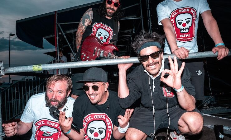 Fünf Männer posieren fröhlich auf einer Bühne, tragen T-Shirts mit Totenkopf-Logo und lächeln in die Kamera. Einer hält eine Gitarre., © clubCANN - Stuttgarter Jugendhaus gGmbH Fünf Männer posieren fröhlich auf einer Bühne, tragen T-Shirts mit Totenkopf-Logo und lächeln in die Kamera. Einer hält eine Gitarre., © clubCANN - Stuttgarter Jugendhaus gGmbH