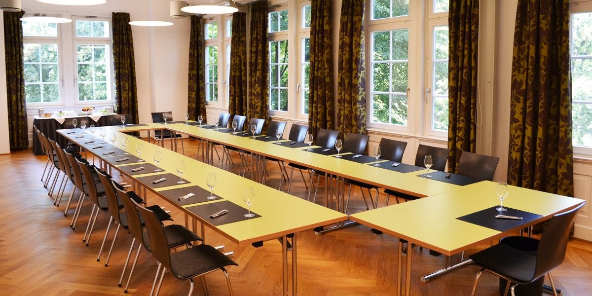 A bright meeting room with yellow U-shaped tables, black chairs and large windows with curtains. Glasses and writing utensils are provided., &copy; ECOINN Hotel am Campus