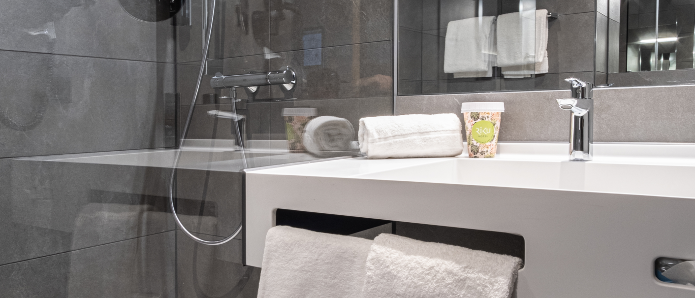 Modernes Badezimmer mit grauen Fliesen, einem wei&szlig;en Waschbecken, Handt&uuml;chern und einem Becher mit Hotel-Logo auf der Ablage., &copy; 13 6 Design GmbH (Judenhof 10, 89073 Ulm)