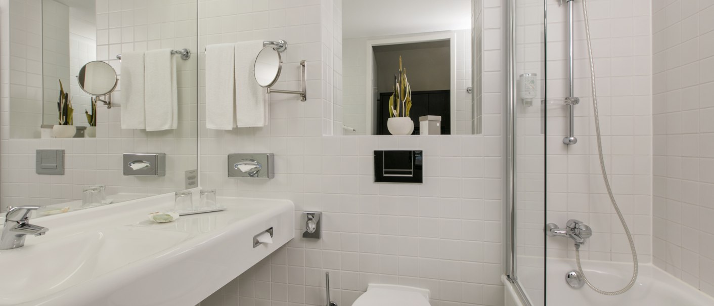 Modernes Badezimmer mit weißen Fliesen, Waschbecken, Toilette, Badewanne und Spiegel. Handtücher und Pflanzen als Dekoration., © ARCOTEL Hotels