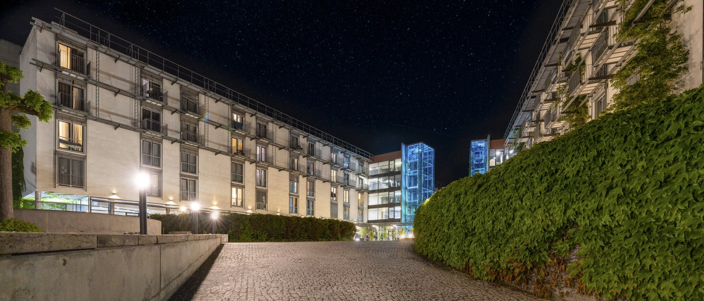 Night view of a modern building complex with illuminated windows, glass elevators and a paved path surrounded by green plants., © PLAZA Hotelgroup GmbH