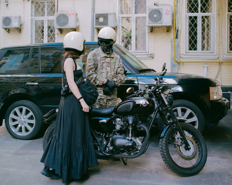 Zwei Personen mit Helmen stehen neben einem Motorrad vor einem schwarzen Auto. Eine Person trägt ein schwarzes Kleid, die andere Tarnkleidung., © Anya Tsaruk Zwei Personen mit Helmen stehen neben einem Motorrad vor einem schwarzen Auto. Eine Person trägt ein schwarzes Kleid, die andere Tarnkleidung., © Anya Tsaruk