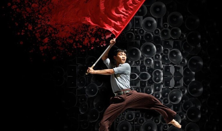 Ein Mann schwingt eine rote Fahne vor einer Wand aus Lautsprechern. Der Hintergrund ist dunkel, die Fahne leuchtet kräftig rot., © Theaterhaus Stuttgart e.V. Ein Mann schwingt eine rote Fahne vor einer Wand aus Lautsprechern. Der Hintergrund ist dunkel, die Fahne leuchtet kräftig rot., © Theaterhaus Stuttgart e.V.