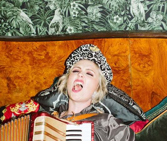 A person in a leopard hat plays an accordion and sings passionately in front of a tropical wallpaper., © Rosenau Kultur e.V. A person in a leopard hat plays an accordion and sings passionately in front of a tropical wallpaper., © Rosenau Kultur e.V.