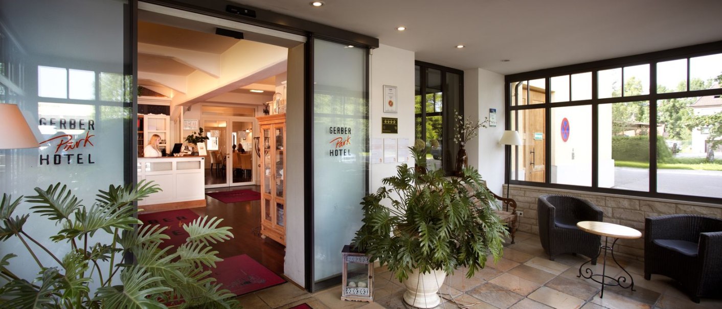Hotel entrance with plants, seating area and reception. Large windows let in plenty of light. The floor is tiled, red carpets lead to the reception., © TOMAS
