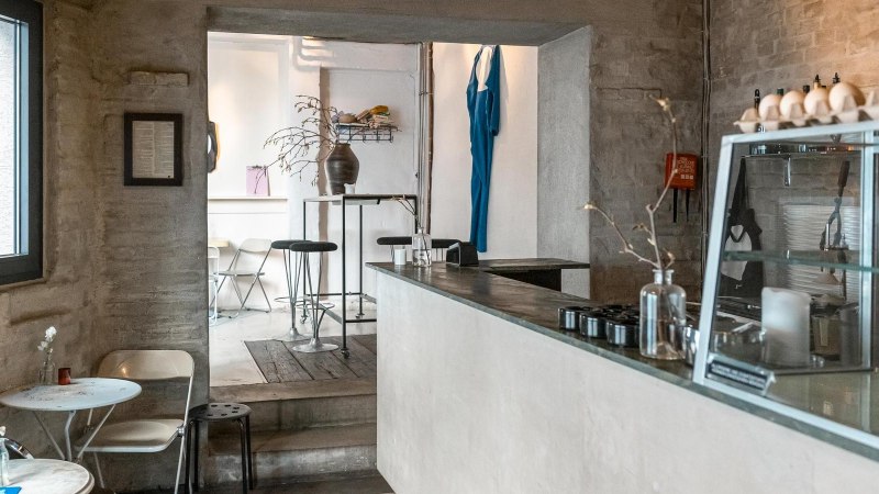 Modernes Café mit minimalistischer Einrichtung, weißen Wänden und Holzmöbeln. Ein blaues Kleidungsstück hängt an der Wand, Pflanzen auf Tischen., © SMG, Sarah Schmid Modernes Café mit minimalistischer Einrichtung, weißen Wänden und Holzmöbeln. Ein blaues Kleidungsstück hängt an der Wand, Pflanzen auf Tischen., © SMG, Sarah Schmid