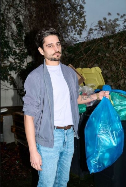 Ein Mann in Jeans und grauer Jacke h&auml;lt einen blauen M&uuml;llsack vor M&uuml;lltonnen. Der Hintergrund zeigt eine bewachsene Wand., &copy; Rosenau Kultur e.V.