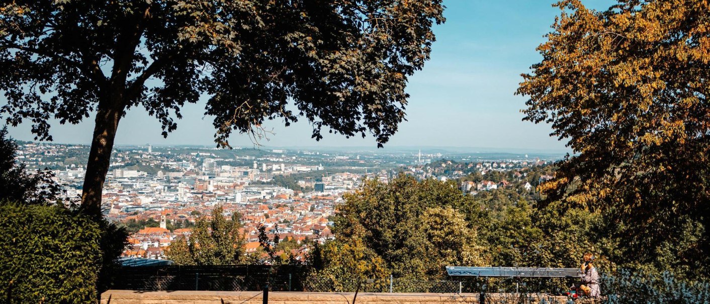 Panoramablick auf eine Stadt mit Bäumen im Vordergrund, die den Blick einrahmen. Eine Person sitzt auf einer Bank und genießt die Aussicht., © Stuttgart-Marketing GmbH, Sarah Schmid