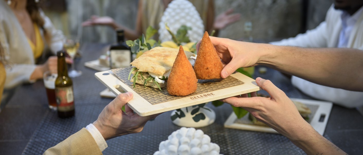 Two people pass a wooden board with arancini and a sandwich at a table with several guests., © CELEBRE