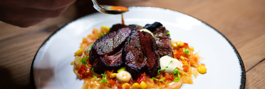 An artfully arranged dish with meat, vegetables and sauce on a white plate. One hand holds a spoon over the plate., &copy; Cara Schmid