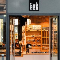 Eingang einer Bäckerei mit dem Schild 'Zeit für Brot'. Innen sind Regale mit Brot und Gebäck zu sehen., © Zeit für Brot Stuttgart Eingang einer Bäckerei mit dem Schild 'Zeit für Brot'. Innen sind Regale mit Brot und Gebäck zu sehen., © Zeit für Brot Stuttgart