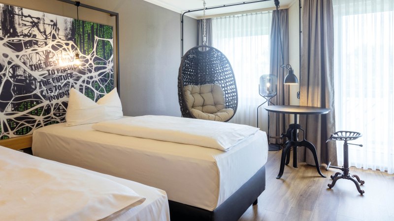 Modernes Hotelzimmer mit zwei Einzelbetten, einem Hängesessel, einem runden Tisch und einem Hocker. Große Fenster mit Vorhängen lassen viel Licht herein., © Seminaris Hotel Bad Boll Modernes Hotelzimmer mit zwei Einzelbetten, einem Hängesessel, einem runden Tisch und einem Hocker. Große Fenster mit Vorhängen lassen viel Licht herein., © Seminaris Hotel Bad Boll