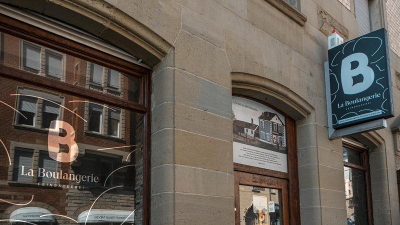 Außenansicht der Bäckerei 'La Boulangerie' mit Schaufenster und Schild. Das Gebäude hat eine steinerne Fassade und spiegelt umliegende Architektur wider., © SMG, Sarah Schmid
