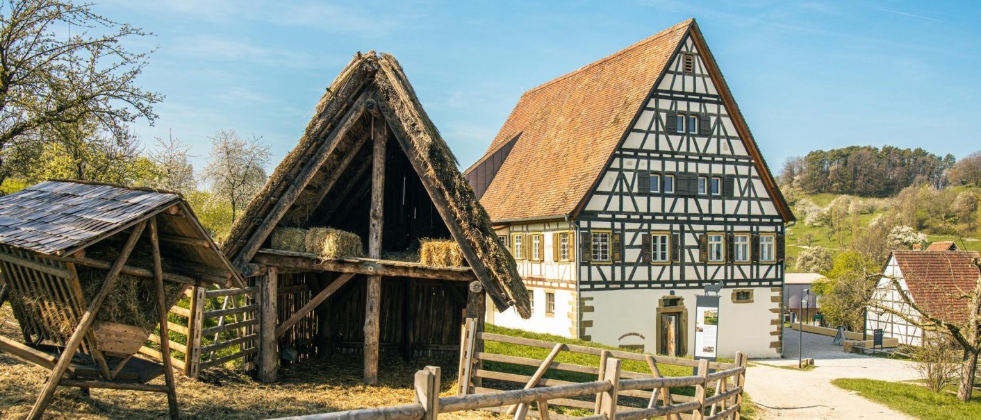 Half-timbered house and barn in the Beuren open-air museum, surrounded by blossoming trees and green hills under a blue sky., &copy; Stuttgart-Marketing GmbH, Sarah Schmid