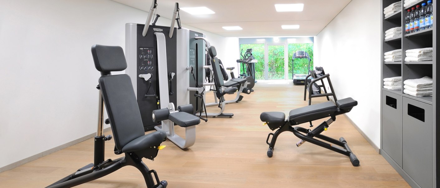 A modern fitness room with various exercise machines, weight benches and a shelf with towels and water bottles., &copy; Schlosshotel Monrepos