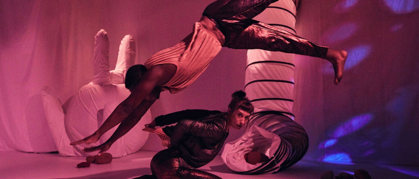 Zwei Akrobaten in einem pink beleuchteten Raum. Einer springt elegant über den anderen, der in einer gebückten Position verharrt., © JES, Alex Wunsch Zwei Akrobaten in einem pink beleuchteten Raum. Einer springt elegant über den anderen, der in einer gebückten Position verharrt., © JES, Alex Wunsch