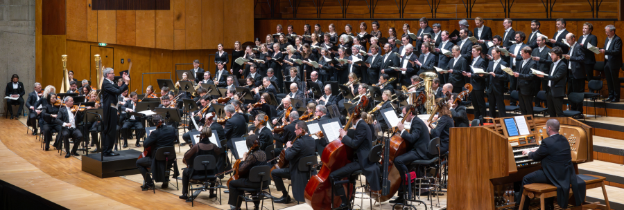 A large orchestra and choir perform in a concert hall. The conductor stands in front of the musicians while an organist plays the organ., &copy; Holger Schneider